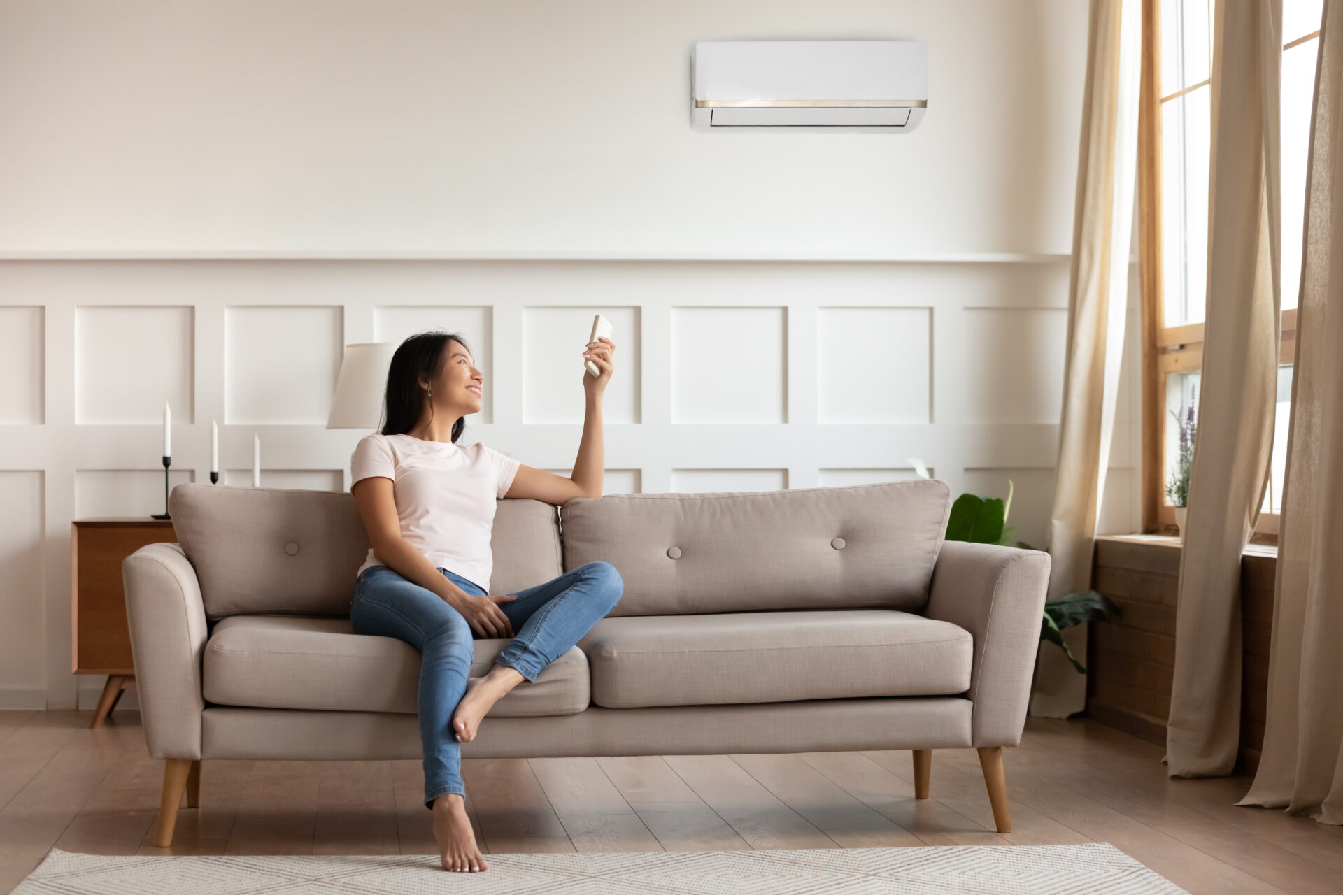 Young asian woman switching on air conditioner while resting seated on couch in modern interior living room, holds remote climate control cooler system manage temperature at contemporary home concept